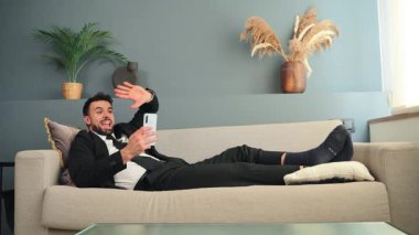 Akıllı takım elbiseli genç bir adam, akıllı telefonunu kullanarak bir video çağrısına cevap verirken koltukta rahatça uzanır. Sohbeti sırasında rahat ama profesyonel bir tavır sergiler..