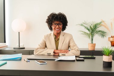 Kendine güvenen bir iş kadını önemli belge ve notları incelerken masasına gülümsüyor. Mesleki giyimi ve misafirperver tavrı olumlu bir atmosfer yaratıyor.