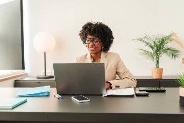 Takım elbise giymiş kararlı ve odaklanmış bir Afro-Amerikan kadını. Ofis gereksinimleri ve bitkilerle çevriliyken bilgisayarında önemli bir işle meşgul..