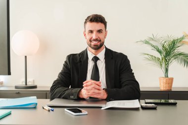 Masasında otururken gülümseyen, resmi bir takım elbise giymiş profesyonel bir toplantıya hazır kendinden emin bir işadamı. Cana yakın, odaklanmış, profesyonellik ve çekicilik yayıyor..