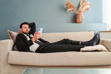 Genç bir iş adamı rahat bir şekilde kanepede dinlenirken akıllı telefonunu çeşitli faaliyetlerde bulunmak için kullanıyor. Ev ortamında rahatlama ve üretkenliğin bir karışımını yansıtıyor..