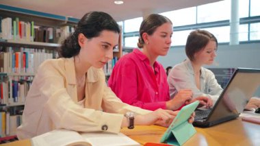 Üç kendini adamış kız öğrenci, bir kütüphane ortamında akademik başarılarını ve üretkenliklerini artırmak için dijital cihazlardan ve kitaplardan yararlanan işbirlikçi bir çalışma oturumuna katıldılar..