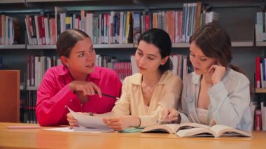 İşbirlikçi bir çalışma oturumuna katılan üç özenli öğrenci, fikir alışverişinde bulunuyor ve akademik anlayış ve takım çalışması becerilerini geliştirirken bir kütüphanede notlarını gözden geçiriyorlar..
