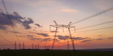 Tarım alanındaki yüksek voltaj hatları ve güç pilonları doymuş günbatımı gökyüzüne karşı.
