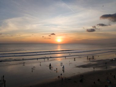 Akşam kumsalında siluetli figürler, uzak güneş yansıtıcı kıyı şeridinde sörfçülerle birlikte batıyor.