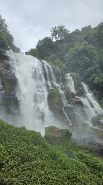Nefes kesen bir şelale bulutlu bir gökyüzünün altında canlı yeşilliklerle çevrili engebeli kayalardan zarifçe dökülüyor. 