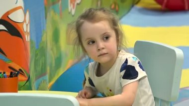 Küçük sarışın kız etrafına bakarak oyun odasında çizer