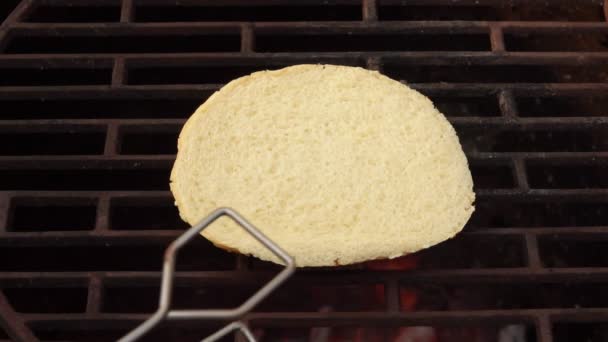 Gros plan du pain burger retourné sur la grille du gril avec pinces de cuisine — Video