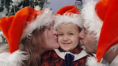 Noel Baba şapkalı mutlu ebeveynler Noel ağacının altında küçük kızlarını öpüyor.