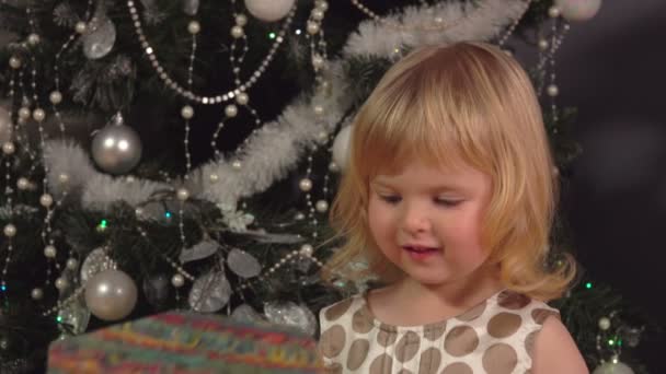 Een schattig klein meisje zit onder de kerstboom en opent cadeautjes — Stockvideo