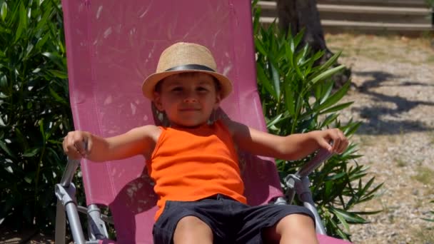 O menino alegre em um chapéu de palha está deitado em uma cadeira de praia rosa — Vídeo de Stock