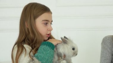 Uzun saçlı örgü elbiseli kız sevimli, gri tüylü küçük bir tavşanı okşuyor. 