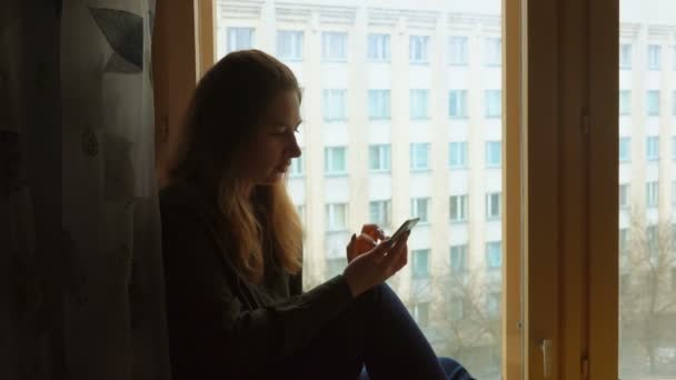 Una giovane donna dai capelli lunghi sta usando un telefono cellulare seduto vicino alla grande finestra — Video Stock