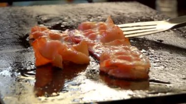 Düz ızgarada metal maşayla çevrilmiş ince domuz pastırması şeritleri.