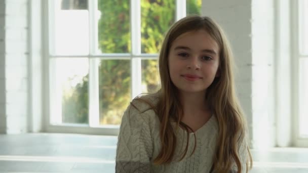 Красива маленька довгошерста дівчинка посміхається і сидить у білому сонячному залі — стокове відео