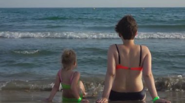 Anne ve kızı sırtı kameraya dönük bir şekilde sahilde oturuyorlar. 