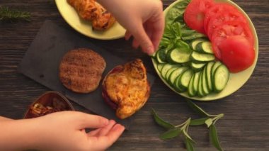 Ellerin üst görüntüsü hamburgerin ekmeğine ızgara tavuk filetosu koyuyor.