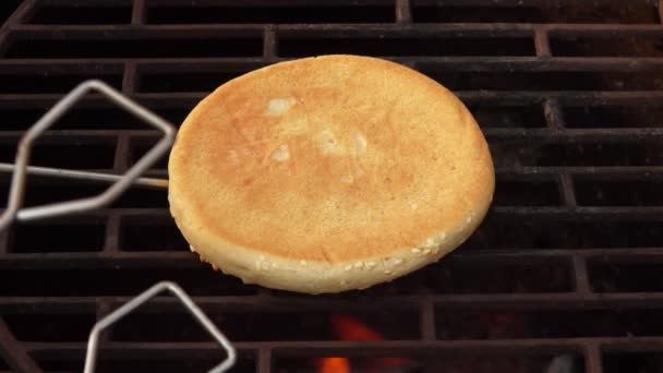Gros plan du pain burger au sésame retourné sur la grille du gril au-dessus du feu ouvert — Video