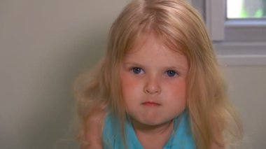 Sarı saçlı küçük bir kız dudaklarını ısırıyor ve kameraya bakıyor.