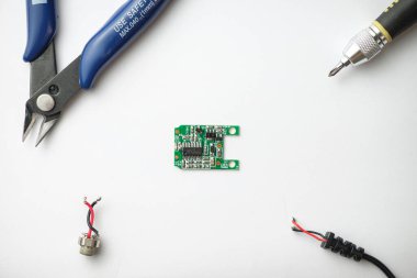 Temiz bir çalışma alanında, küçük yeşil bir devre kartı pense ve tornavida da dahil olmak üzere aletlerin arasında duruyor. Kablolar ve konnektörler yakınlarda, bir elektronik projenin başladığına işaret ediyorlar..