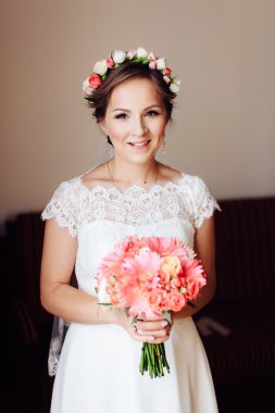 Cute lovely cheerful young bride in beautiful wreath of flowers showing bouquet.