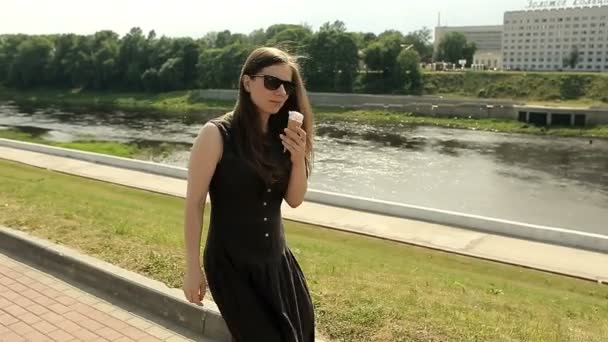 Hezká dívka v sluneční brýle na ulici chůzi a jíst zmrzlinu. Stabilní cam — Stock video