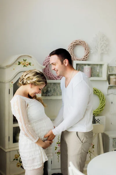 Feliz embarazada esposa con marido vestido en blanco ropa hermosa tocando vientre en casa . — Foto de Stock