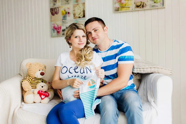 Jovem grávida e marido no sofá branco no quarto com roupas de bebê . Imagem De Stock