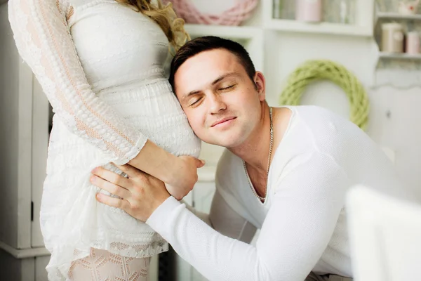 Ευτυχισμένη σύζυγος με κλειστά μάτια, ακούγοντας την κοιλιά έγκυος γυναίκα του στο σπίτι Εικόνα Αρχείου