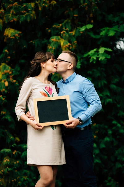 Förväntar sig gravida lyckliga snygga paret håller en tom träkol styrelse. — Stockfoto