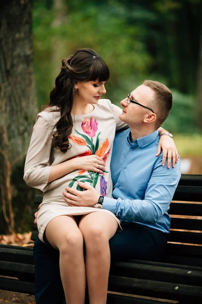 Όμορφη έγκυος κομψό ζευγάρι χαλαρωτικό εκτός στο φθινοπωρινό πάρκο κάθεται σε παγκάκι. Εικόνα Αρχείου