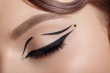 Kapalı bir kadın gözünün makrosu siyah kanatlı göz kalemi ve küçük altın nokta süslemesi, yumuşak ışıklandırma kirpik eğrisi ve editoryal güzellik kullanımı için göz kapağı süslemesi.