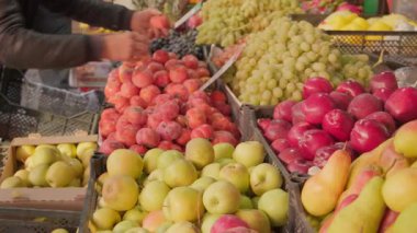 Taze üzümler, elmalar ve hurmalar açık hava pazarında ön planda sergilenirken, arkada bulanık bir muz alımı sahnesi görülüyor. Günlük yaşam tarzı, rutin alışveriş ve yerel toplum kültürü.