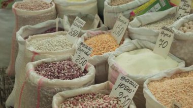 Alışveriş yapan el, açık fasulye çuvallarına uzanıyor, el yazısı fiyat etiketleri, el yapımı pullar ve mısırlar, toplu gıda ürünleri ve renk kontrastlı gizli pazar tezgahı sahnesi. Yerel pazar ticareti