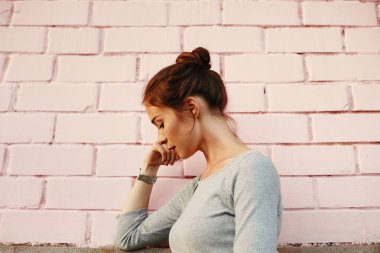 Pembe Duvara yaslanan genç bir kadın, dağınık kollu gri süveter ve bilek saati olan düşünceli bir öğrenci portresi, yumuşak doğal ışık, sessiz pastel tonları, desenli tuğla fon, aşağı doğru bakan bir bakış.