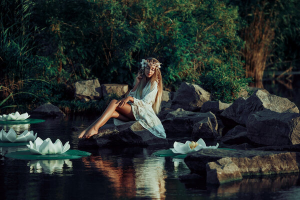 Fairy sitting on a rock in the water.Mysterious lake with big lilies. Background fairy forest and river swamp. A wreath made of shells, handmade. Fashionable toning.