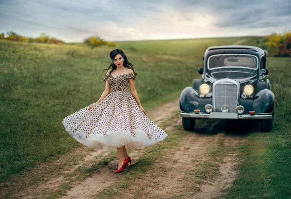 Jonge mooie vrolijke retro vrouw bij de oude auto. Aantrekkelijke elegante dame in vintage witte polka zwarte stip retro jurk. Pin-up kapsel. Vrolijk meisje op de weg. Herfst natuur, gele bomen — Stockfoto