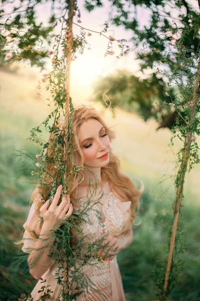 Jovem mulher loira feliz na imagem de uma ninfa da floresta em um vestido vintage pêssego leve, fica perto de uma árvore. Menina modelo de moda posando no fundo do verão natureza verde, sol pôr do sol. Cabelo loiro longo. — Fotografia de Stock