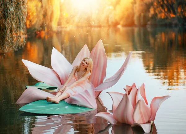 Glad fantasi ung blond kvinna liten älva prinsessa sitter i rosa lotus blomma på sjön vatten. Alvflicka, med leende ansikte. Hösten natur bakgrund apelsin träd gudomlig magi sol ljus. Rosa klänning — Stockfoto