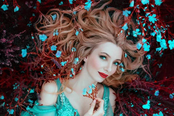 Bella donna elfo fantasia felice con i capelli biondi si trova sogni su erba, prato fiorito, fiori blu. Ritratto viso sorridente, labbra rosse trucco elegante. Ragazza romantica principessa a riposo godendo la vita — Foto Stock