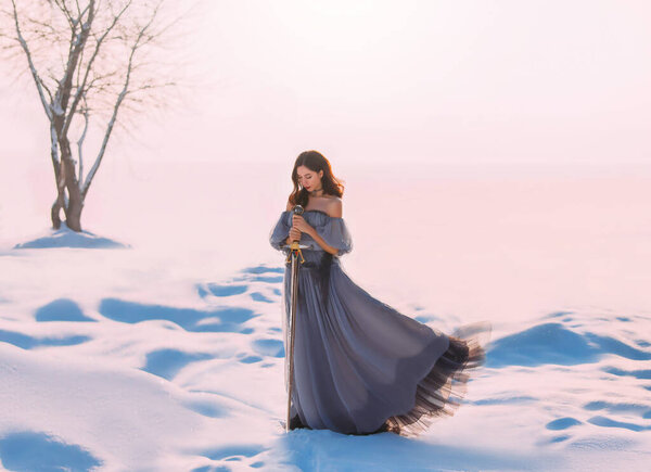Женщина-фантазия воительница принцессы, держащая в руках средневековый железный меч. Снежная королева сказки. Винтажное серо-голубое платье. Белый зимний фон природы, долина, дерево. Девушка-эльф со стальным лезвием. Фото искусства.
