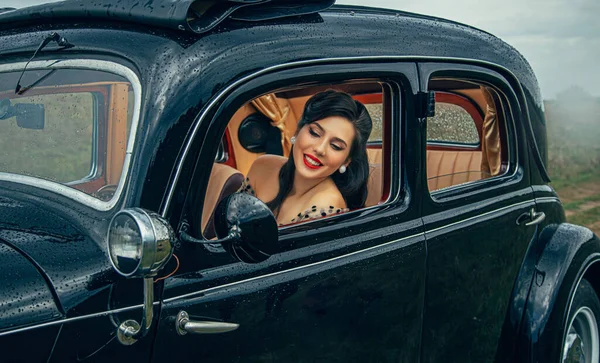 Joyeux jeune femme rétro assis dans une voiture noire au volant et regarde par la fenêtre avec le sourire sur son visage. Belle fille pilote en robe vintage, coiffure pin-up, maquillage des lèvres rouges. Brume de pluie mauvais temps — Photo