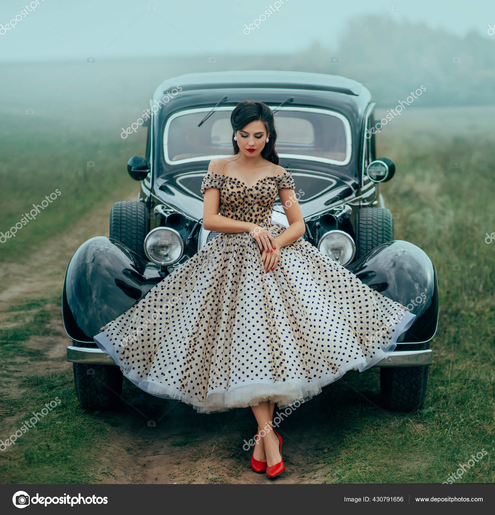 Young beautiful sexy woman in pin-up style clothes posing near black retro  car. Polka dot white dress, vintage hairstyle, red high heels. Background  road green nature fog. Girl fashion model driver –