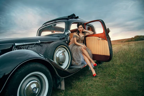 Jeune belle femme sexy dans des vêtements de style pin-up est assis posant en voiture rétro noire. Robe blanche à pois, coiffure vintage, talons hauts rouges. Contexte route nature été. fille modèle de mode pilote — Photo