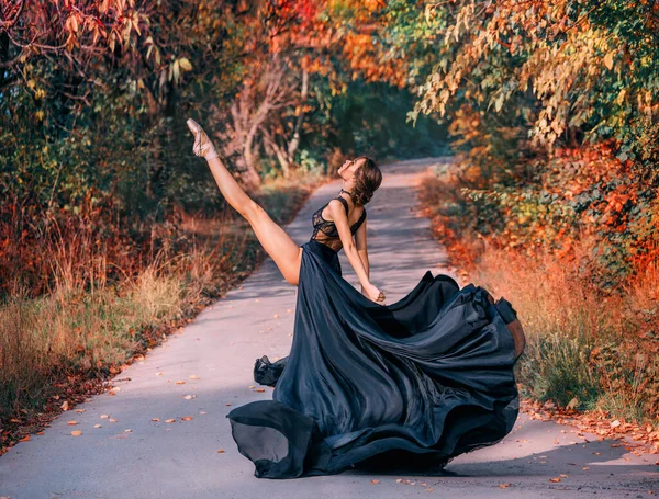 Fantasie Schöne Ballerina, die auf der Straße tanzt. Herbst Natur Hintergrund Bäume. Modemodel posiert in Bewegung. Langes seidenes schwarzes Kleid, Rock fliegt im Wind. Frauenballett-Tänzerin Bein wird gespalten — Stockfoto