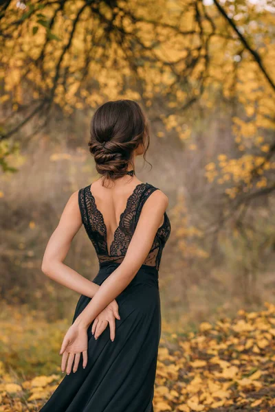 Fantastische vrouw in een zwarte kanten jurk. kale open sexy rug tegen de achtergrond van een herfstbos. Elegant vintage middeleeuws kapsel voor lang haar. Fashion prinses model poseren. gezond donker haar — Stockfoto