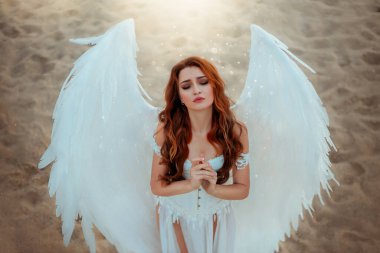 Beyaz sahte kuş kanatlı ve yapay tüylü melek kostümlü fantezi kadın. Elleri dua ederken katlanmış, gözleri kapalı tanrıça kız. Doğa sahili, kum çölü kutsal güneş ışığı.