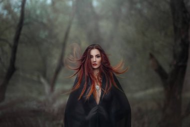 Esrarengiz fantezi gotik kadın. Kötülüğe takıntılı kara cadı. Siyah pelerinli kızıl saçlı şeytan kız. Kızıl saçlar rüzgarda dalgalanıyor. Karanlık, sık ormanın arka planı, ağaçlar. Gözlerde skleral lensler