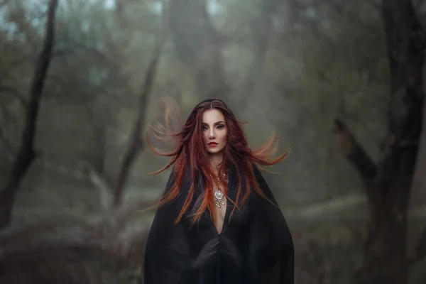 Misteriosa fantasia mulher gótica bruxa negra obcecada pelo mal. Rapariga de cabelos vermelhos demônio no capuz de capa de vestido preto. O cabelo ruivo treme ao vento. Fundo de floresta profunda densa escura, árvores. Lentes esclerais nos olhos — Fotografia de Stock