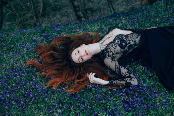 Fantasía mujer pelo rojo acostado sueño sueño. hierba pradera floreciente bosque oscuro. Reina vampiro dama medieval. largo en negro vestido de encaje pecho abierto, sexy ninfa caliente piernas desnudas sexy. luna luz noche magia — Foto de Stock